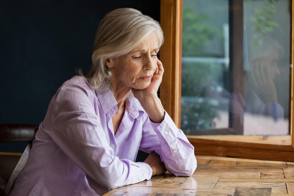 sad senior woman sitting alone a table in front of window